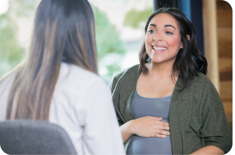 Smiling pregnant woman cradling her belly while talking to another person indoors