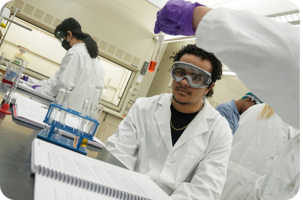 Student in goggles and white lab coat at lab bench with classmates and glassware