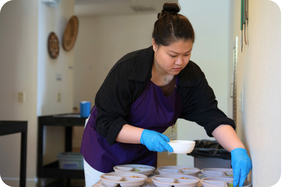 Focused woman in purple apron and blue gloves arranging bowls on a kitchen counter.