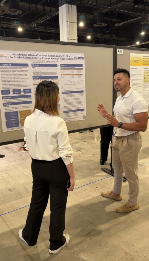 Two colleagues at conference poster: man in white polo gesturing, woman in white blouse listening