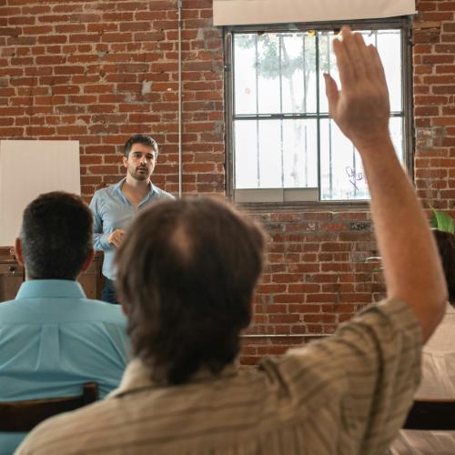 A man speaking to an audience; one person raises their hand to ask a question.