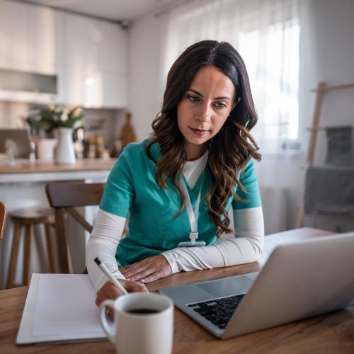 A woman in a teal shirt sitting at a wooden table, working on a laptop with a cup of coffee.